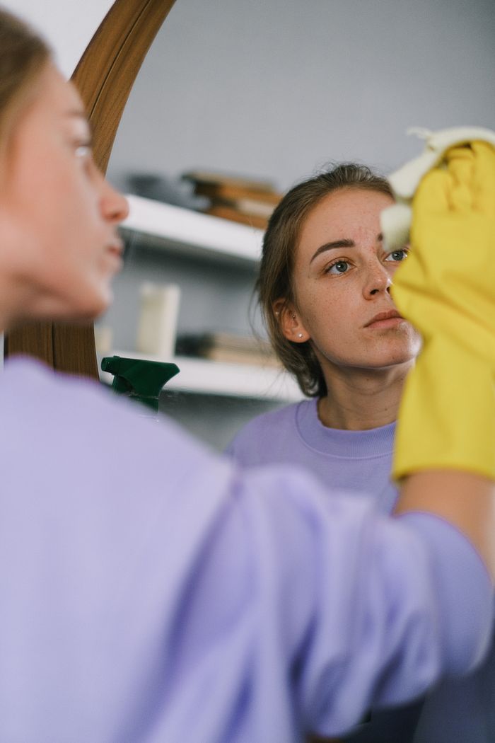 Femme recadrée portant un sweat-shirt violet et des gants en latex, nettoyant un miroir dans un appartement lumineux en journée